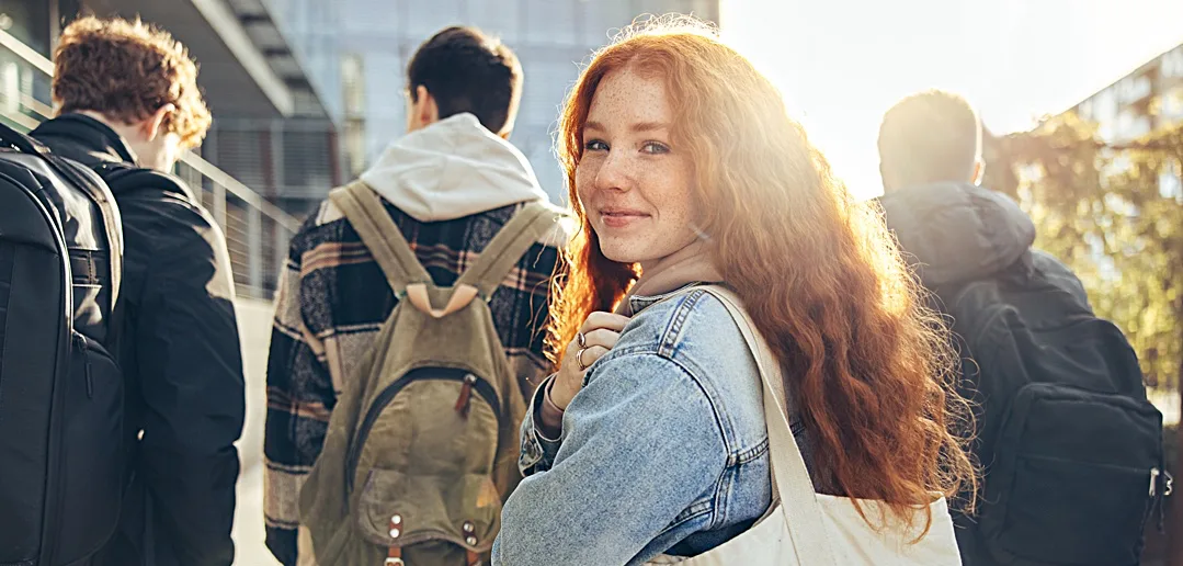 Smiling young woman with red hair walking outdoors with fellow students carrying backpacks.