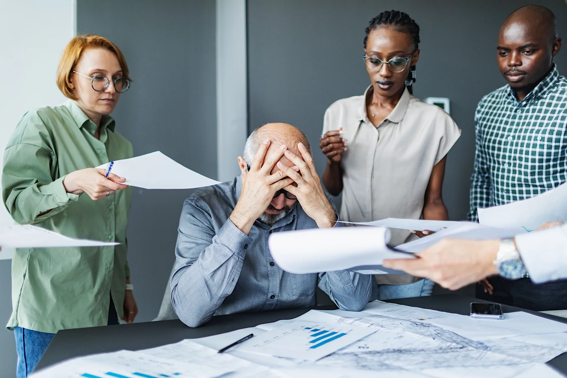 Stressed man at work overwhelmed by colleagues, papers and deadlines in a busy office environment.