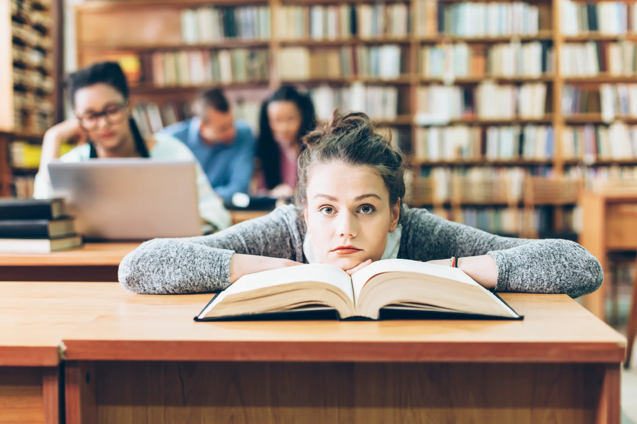 Student in a library looking tired, surrounded by books and peers studying.
