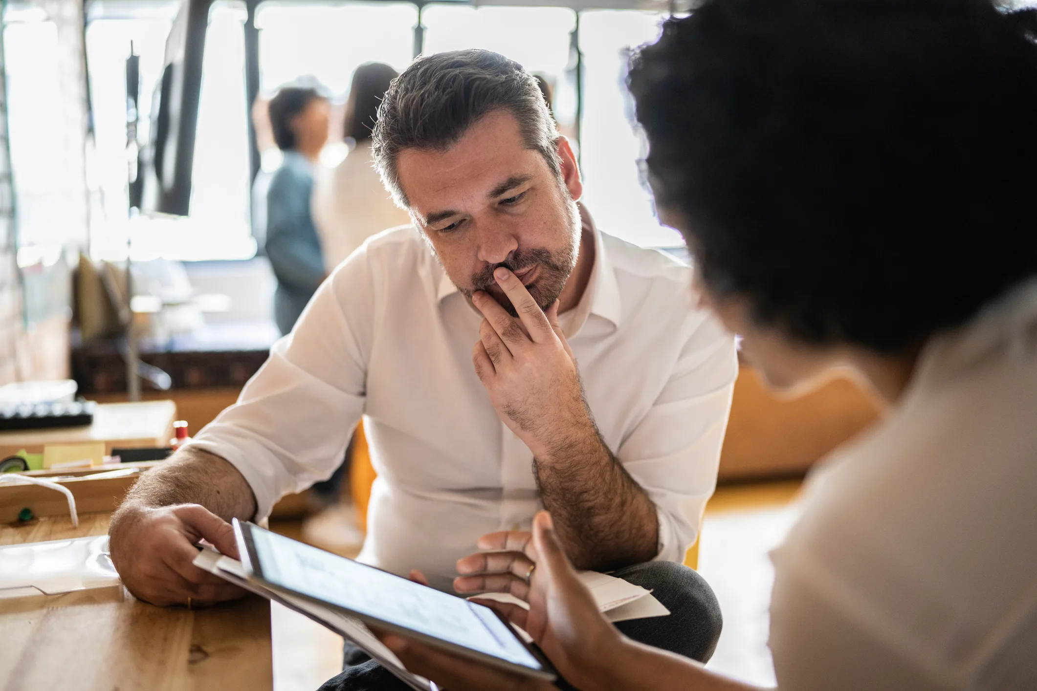 Man in white shirt discussing with colleague, using digital tablet in office setting.