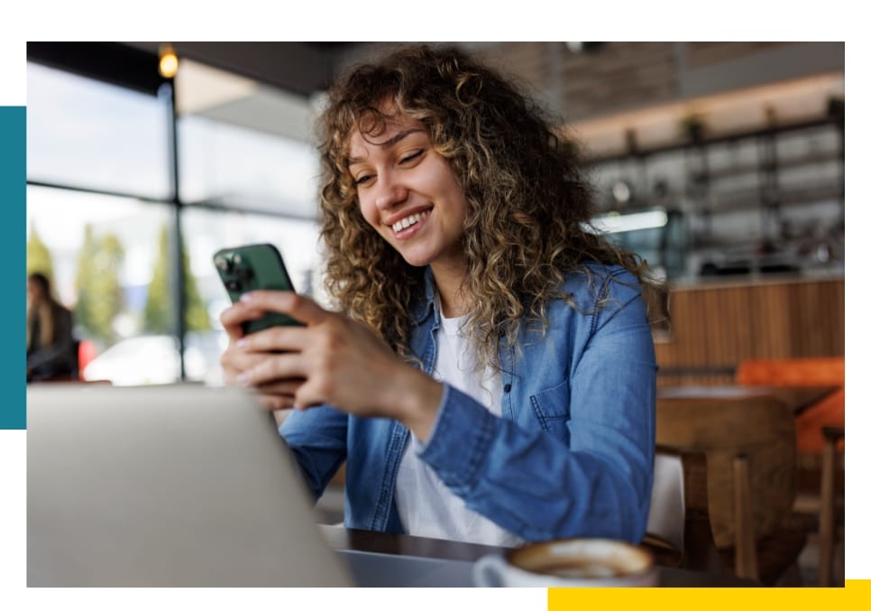 Woman smiling while using a smartphone and laptop in a cozy café setting.