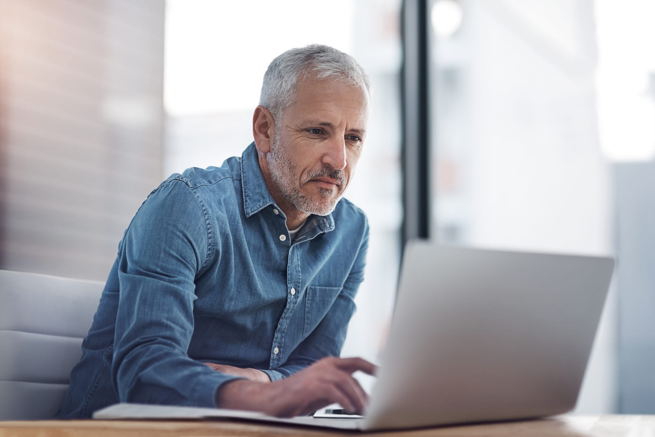 Man in denim shirt focused on work on laptop in bright office setting.