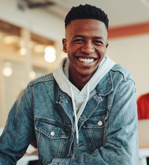 Young Black man with short hair smiling broadly at the camera, wearing a denim jacket over a gray hoodie in a bright indoor space.